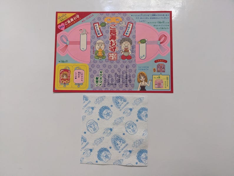 May include: A Japanese souvenir set. The set includes a pink and purple illustrated card with cartoon characters and text, and a white cloth printed with blue cartoon faces.