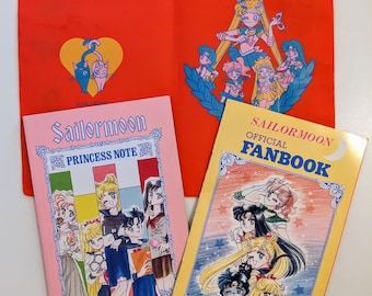 Op de afbeelding: Een roze en gele Sailor Moon Princess Note boek en een gele Sailor Moon Official Fanbook met een rode map. De map heeft een Sailor Moon logo en een afbeelding van de Sailor Scouts. De boeken hebben afbeeldingen van de Sailor Scouts op de omslagen.
