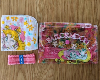 Op de afbeelding: Een roze en witte handdoek met Sailor Moon-thema, een roze lint met het woord "Nakayoshi" erop gedrukt en een doorzichtige roze plastic portemonnee met een Sailor Moon-ontwerp en de tekst "Usagi en Chibiusa" erop gedrukt.