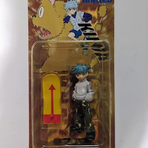 May include: A Hunter x Hunter action figure in a clear plastic package. The figure depicts a character with spiky blue hair, wearing a white shirt and black pants. The packaging features cartoon illustrations and the text "HUNTER x HUNTER".