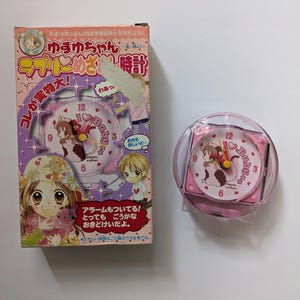 May include: A pink and white alarm clock with a cartoon design, featuring the text "Baby" and Japanese text. The clock is in a clear plastic case, with a matching box. The box has Japanese text and illustrations.