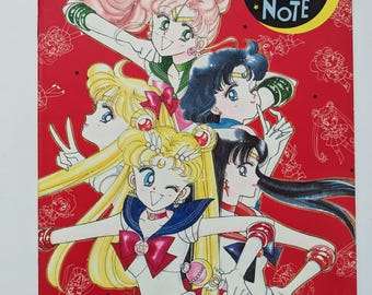 Op de afbeelding: Een rode Sailor Moon Fantasy Note boekomslag met vijf anime-personages in kleurrijke outfits. De omslag heeft de tekst "Sailor Moon Fantasy Note" in een gele cirkel. De achtergrond is rood met kleine witte illustraties.