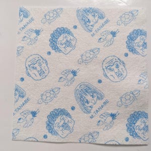 May include: Square white fabric featuring a repeating blue pattern of cartoon faces, turtles, birds, and dots. The text "M. TANABE" is also visible in the design.