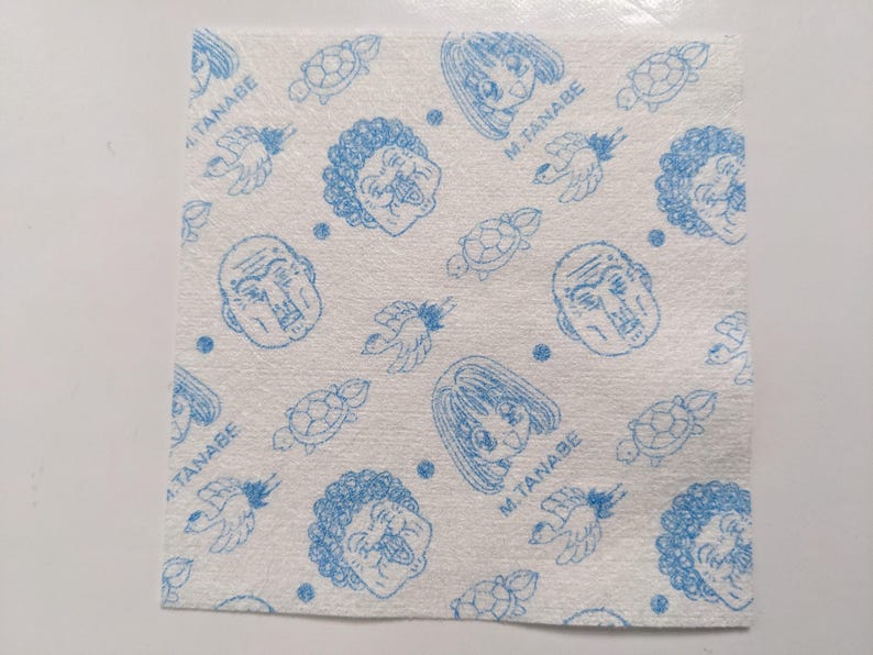 May include: Square white fabric featuring a repeating blue pattern of cartoon faces, turtles, birds, and dots. The text "M. TANABE" is also visible in the design.