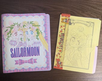 Op de afbeelding: Een roze en paarse map met een Sailor Moon-ontwerp met twee animemeisjes met bloemen in hun haar. De map heeft een ruitpatroon en de tekst "Sailor Moon" in paarse letters. De afbeelding toont ook een geel kleurboek met een zwart-witte lijntekening van een Sailor Moon-personage met de tekst "Sailor Moon" op de omslag.