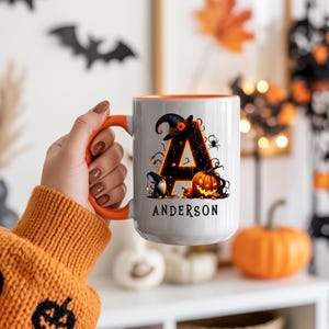 May include: White ceramic mug with an orange handle and interior. The mug features a Halloween-themed design with the letter "A", a witch hat, pumpkins, and the name "Anderson".