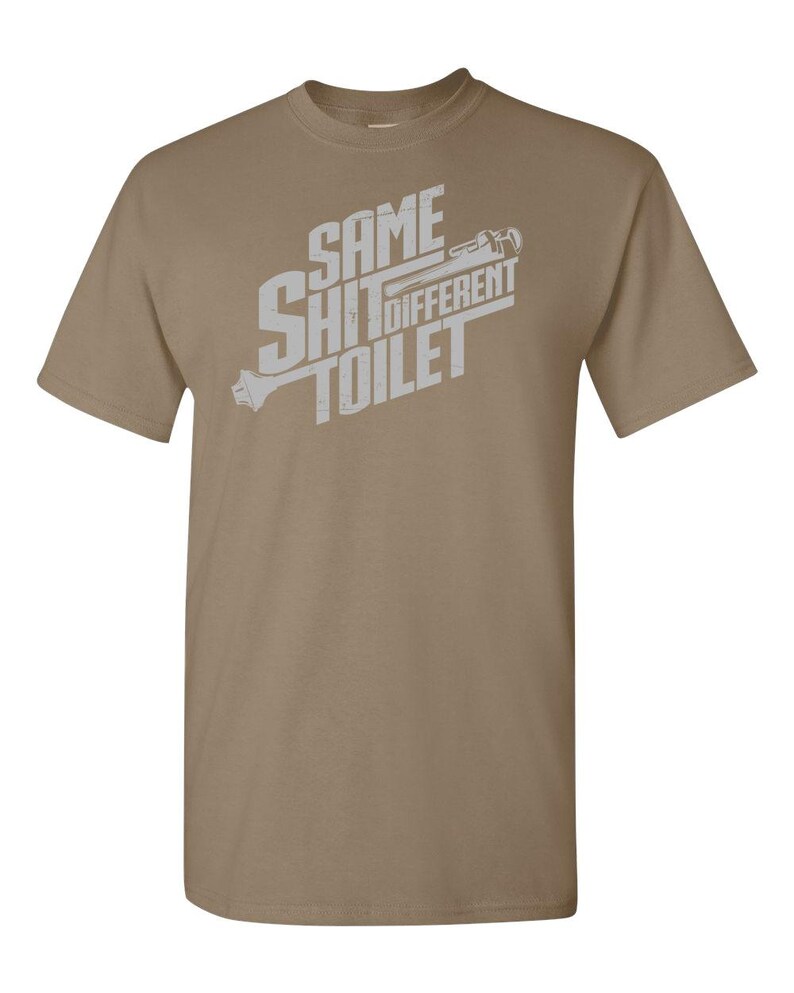 Puede incluir: Una camiseta de color marr&oacute;n claro con un gr&aacute;fico blanco que dice "Same Shit Different Toilet" con una llave inglesa y un gr&aacute;fico de inodoro.