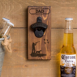 May include: Wooden wall-mounted bottle opener with a black metal opener and the word "DAD" engraved on the top. The opener features a silhouette of a hunter with a dog and birds flying overhead.