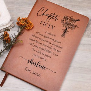 May include: A brown leather journal with the text "Chapter FIFTY" and a floral illustration. The journal includes the name "shirlene" and the year "Est. 2026". A ribbon bookmark is visible.