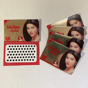 May include: A pack of five black bindi stickers with a sheet of 49 black bindi stickers. The packaging features a gold background with the text "Shilpa Gold" and a woman with a bindi on her forehead.