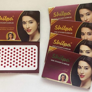 May include: Several boxes of Shilpa Vive Sticker Kumkum, a cosmetic product. The boxes are burgundy with gold lettering and feature a woman's image. One box is open, revealing a sheet of small red dots.