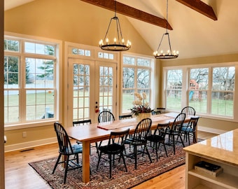 May include: A dining room with a large wooden table and chairs. The table is set with a white tablecloth and a centerpiece of flowers. The room has large windows that let in natural light. The walls are painted a light beige color. There are two black chandeliers hanging from the ceiling.
