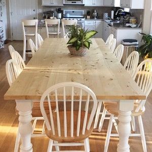 May include: A large, rectangular, wooden table with a natural finish and white painted legs. The table is surrounded by white chairs with a wood seat. A green plant sits in a pot on the table.