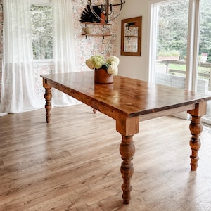 May include: A large, rectangular wooden dining table with turned legs. The table is made of a warm-toned wood and is the focal point of a bright, airy dining room. A vase of white flowers sits on the table. The room features floral wallpaper and sheer curtains.