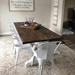 May include: A rustic wooden dining table with a white X-shaped base, surrounded by six white metal chairs. The table is set with a wooden tray, a red and white floral arrangement, and a decorative wooden box. A sign above the table reads "If you want to bring happiness to the whole world, go home and love your family."