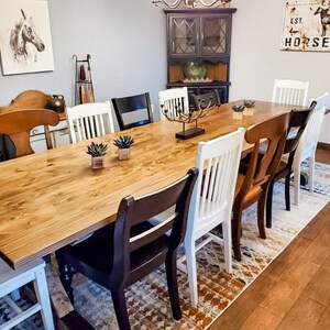 May include: A wooden dining table with a light brown finish, surrounded by chairs in various styles and colors. The table is set with a centerpiece and a few small plants. The room has a light blue wall and a dark wood cabinet with glass doors.