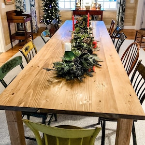 May include: A long wooden dining table with a natural finish is set for a holiday dinner. The table is decorated with a green garland with pine cones and red candles. There are mismatched chairs around the table, some with a green painted finish and others with a natural wood finish.