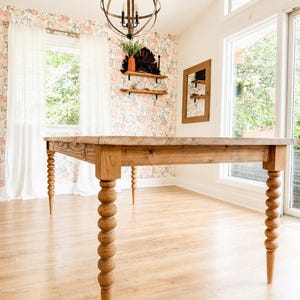 May include: A rectangular wooden dining table featuring four distinct turned legs with a stacked ball design. The table has a natural light brown wood grain finish and a smooth, flat top surface, standing on a light-colored wood floor.