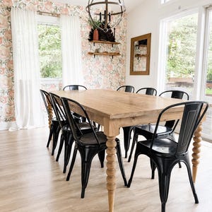 May include: A rectangular wooden dining table with four turned, spindle-style legs, surrounded by six black metal chairs. The table features a natural wood grain finish, while the chairs have a curved backrest and a simple, industrial design.