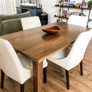 May include: A rectangular dark brown wooden dining table with four legs and a smooth surface. Centered on the table is a small, teardrop-shaped wooden bowl. Four chairs with off-white fabric upholstery and dark brown wooden legs surround the table.