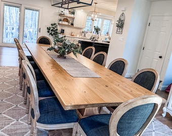May include: A long wooden dining table with a natural finish and a light brown rug underneath. The table is surrounded by chairs with dark blue fabric seats and light wood frames. A black chandelier hangs above the table.