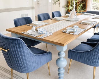 Könnte beinhalten: Ein Esstisch mit einer naturfarbenen Holzplatte und hellblauen gedrechselten Beinen, umgeben von blauen Samtstühlen mit vertikaler Steppung und goldfarbenen Metallbeinen. Der Tisch ist mit weißen Tellern, Platzsets und einem Tischläufer gedeckt.