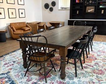 Könnte beinhalten: Ein rechteckiger Esstisch aus dunklem Holz mit gedrechselten Beinen, umgeben von sechs schwarzen Stühlen mit Spindelrücken. Der Tisch steht auf einem großen Teppich mit Blumenmuster in Blau-, Rosa- und Cremetönen in einem Raum mit dunklen Holzbalken.