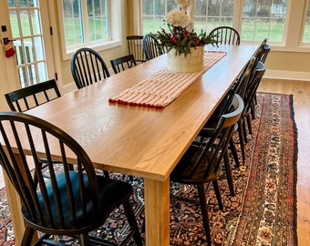 May include: A long rectangular light wood dining table with eight black spindle-back chairs. A white striped table runner with red pom-pom trim sits in the center, topped with a white planter containing white flowers and red berries.