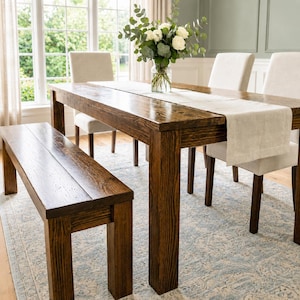 May include: A dark brown wooden dining table with a matching bench and three upholstered chairs. A glass vase filled with white roses and eucalyptus leaves sits on a light beige table runner atop the table, which rests on a patterned rug.