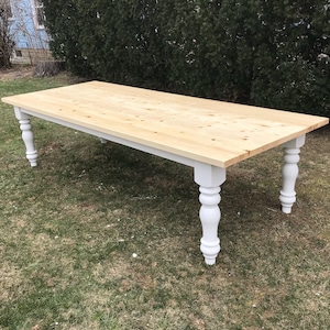 May include: A long, rectangular wooden table with a natural wood finish and white turned legs. The table is sitting on a green grassy lawn.