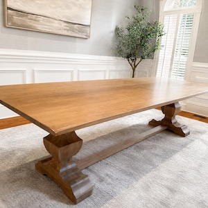 May include: A large rectangular dining table crafted from light brown wood. The table features a smooth, flat tabletop surface supported by two substantial pedestal bases connected by a central horizontal stretcher beam along the floor.