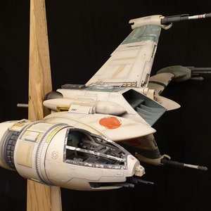 Puede incluir: Un modelo blanco y azul de un caza estelar V-4X-D del universo de Star Wars. El modelo está detallado con una cabina, alas y motores. Está montado en un soporte de madera.