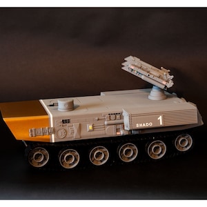 May include: A grey and brown model of a military vehicle with a missile launcher on top. The vehicle has a white stripe along the side and the text "SHADO 1" on the side.