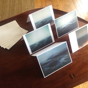 Puede incluir: Cinco tarjetas de felicitación en blanco con una imagen de paisaje marino azul y blanco impresa en cada tarjeta. Las tarjetas están dispuestas sobre una mesa de madera.