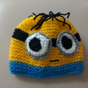 May include: A yellow knitted hat with a blue brim, featuring a black and white eye design and black hair on top. The hat is designed to look like a Minion character.