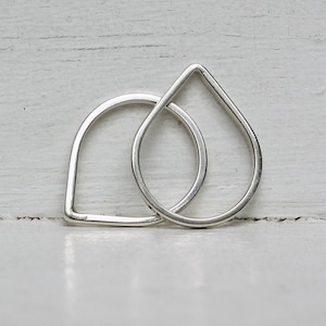 May include: Two silver teardrop-shaped rings, one slightly larger than the other, are stacked on top of each other. The rings are simple and minimalist in design.