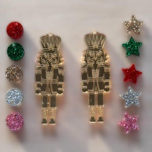 Könnte beinhalten: Zwei goldene Nussknacker-Anhänger mit Glitzer und roten, grünen, goldenen, silbernen und rosafarbenen Sternen-Anhängern mit Glitzer.