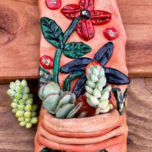 May include: A terracotta-colored ceramic wall planter with a relief design of succulents and flowers. The planter features a variety of green and red accents, including a large red flower and several small red berries. A small succulent plant is next to the planter.