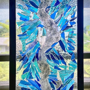 May include: A rectangular stained glass panel with a black frame. The artwork features a central silver-colored design with blue and teal glass pieces arranged to resemble wings or abstract shapes. The glass has a textured appearance.