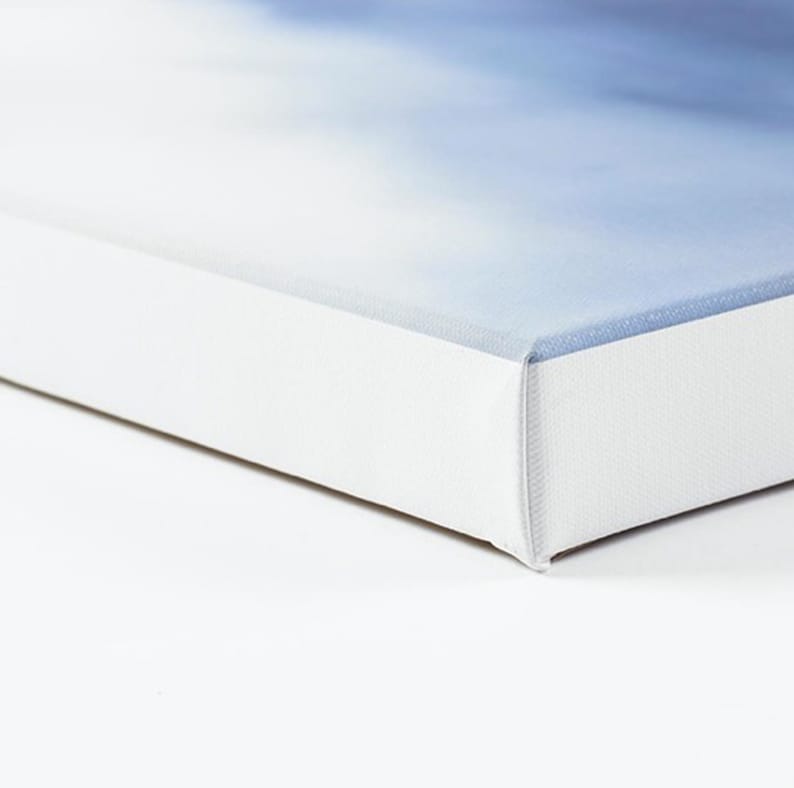 May include: A close-up of a canvas art print with a soft, blurred blue and white abstract design. The canvas is stretched over a white wooden frame, with the edges of the frame visible. The artwork is displayed against a white background.