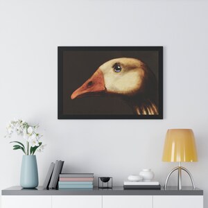 May include: Framed art print featuring a detailed illustration of a goose's head in profile. The artwork showcases the goose's eye, beak, and feathers with a focus on color and texture. The goose has a beige head and a reddish-orange beak. The artwork is framed in a black frame.