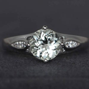 May include: A silver ring featuring a large, round, pale green gemstone. The band has small, clear accent stones on either side of the main stone. The ring is set against a dark grey background.