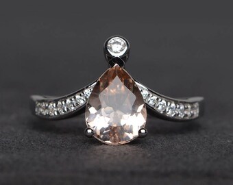 May include: A pear-shaped, peach-colored gemstone ring with a halo of small diamonds and a single diamond accent. The ring has a silver band with additional small diamonds set into the sides.