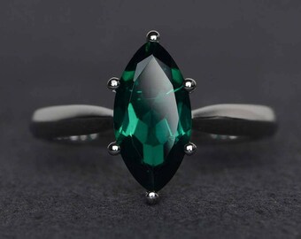 May include: A silver ring featuring a marquise-cut, emerald-green gemstone. The gemstone is set with small silver prongs. The ring band is a simple, polished silver. The ring is set against a dark gray background.