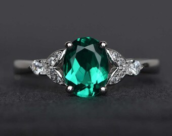 May include: A silver ring with a large, oval emerald green gemstone at its centre. The ring is embellished with smaller, clear gemstones on either side of the main stone, arranged in a decorative design.