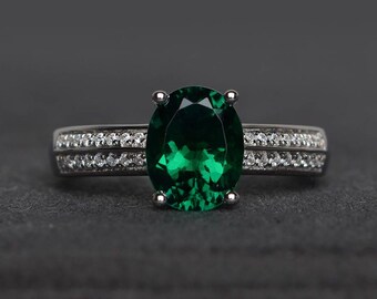 May include: A silver ring with a large, oval emerald. The band has two rows of small, clear gemstones. The ring is set against a dark grey background, emphasising the emerald's vibrant green colour. A piece of jewellery.