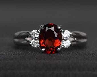 May include: A silver ring featuring a large, oval-cut, deep red gemstone as the centerpiece. Flanking the central stone are smaller, round, clear gemstones. The ring has a sleek, modern design, perfect for a special occasion.