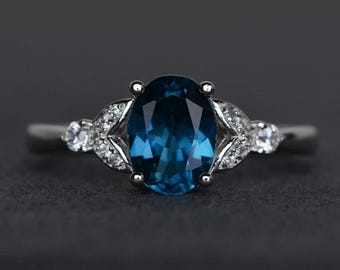 Può includere: Un anello d'argento con una grande pietra preziosa ovale blu scuro al centro. L'anello è impreziosito da pietre preziose più piccole e trasparenti su entrambi i lati della pietra principale, disposte in un motivo decorativo. L'anello è posto su uno sfondo scuro.