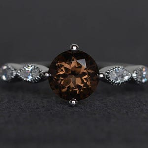 May include: A silver ring featuring a large, round, brown gemstone as the centerpiece. The band is adorned with smaller, clear gemstones on either side of the main stone. The ring is set against a dark gray background.