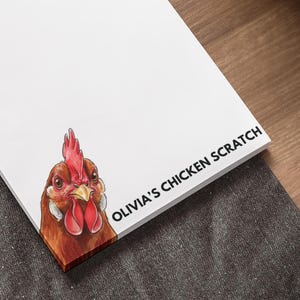 May include: White notepad with a watercolor illustration of a chicken and the text "OLIVIA'S CHICKEN SCRATCH" in black. The notepad is on a dark gray surface next to a laptop and a pen.